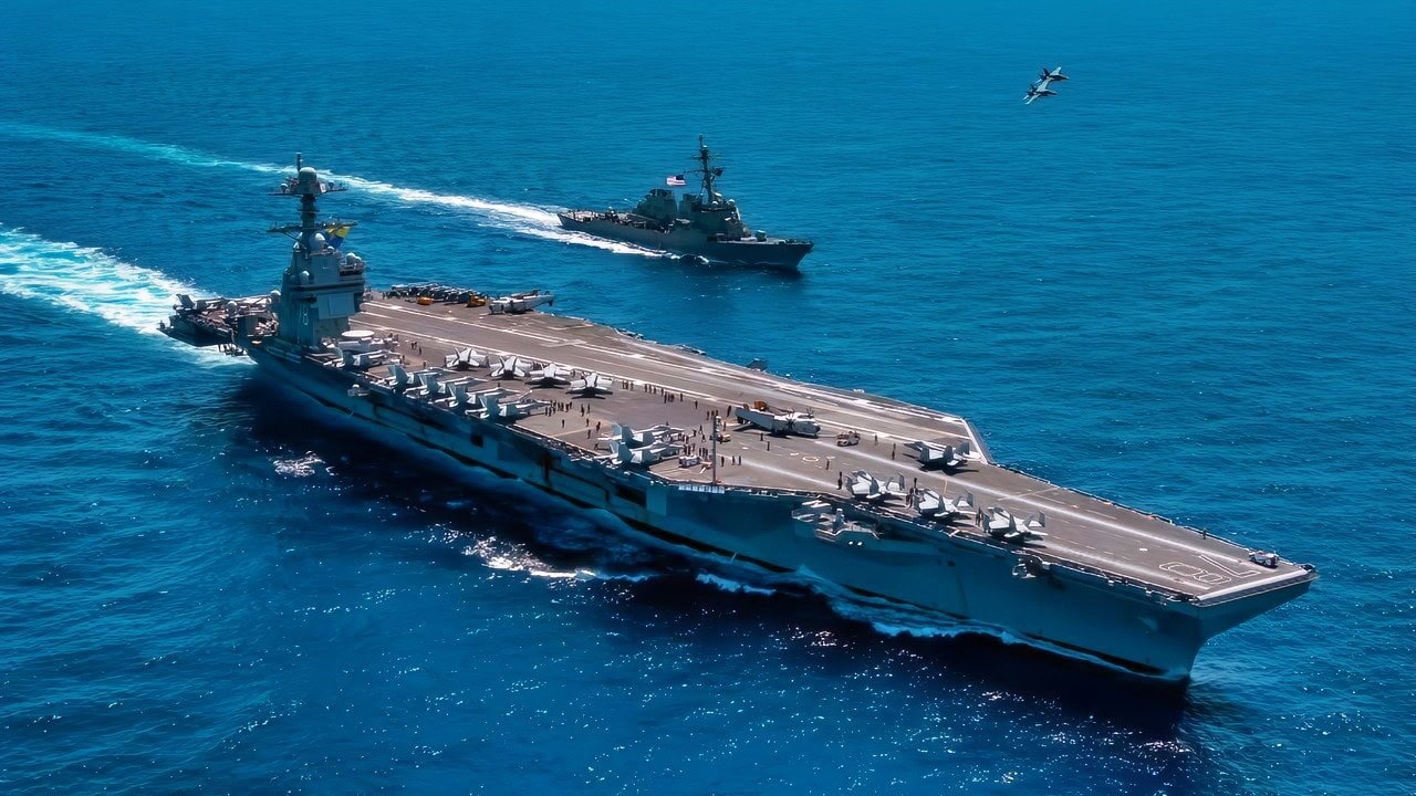 An F/A-18F Super Hornet, attached to the "Blacklions" of Strike Fighter Squadron (VFA) 213 and a F/A-18E Super Hornet, attached to the "Golden Warriors" of Strike Fighter Squadron (VFA) 87 fly over the world's largest aircraft carrier, USS Gerald R. Ford (CVN 78), the Arleigh Burke-class guided-missile destroyer USS Mahan (DDG 72), April 11, 2025. The Gerald R. Ford Carrier Strike Group is underway in the Atlantic Ocean completing integrated naval warfighting training. Composite Training Unit Exercise. Image Credit: Creative Commons.