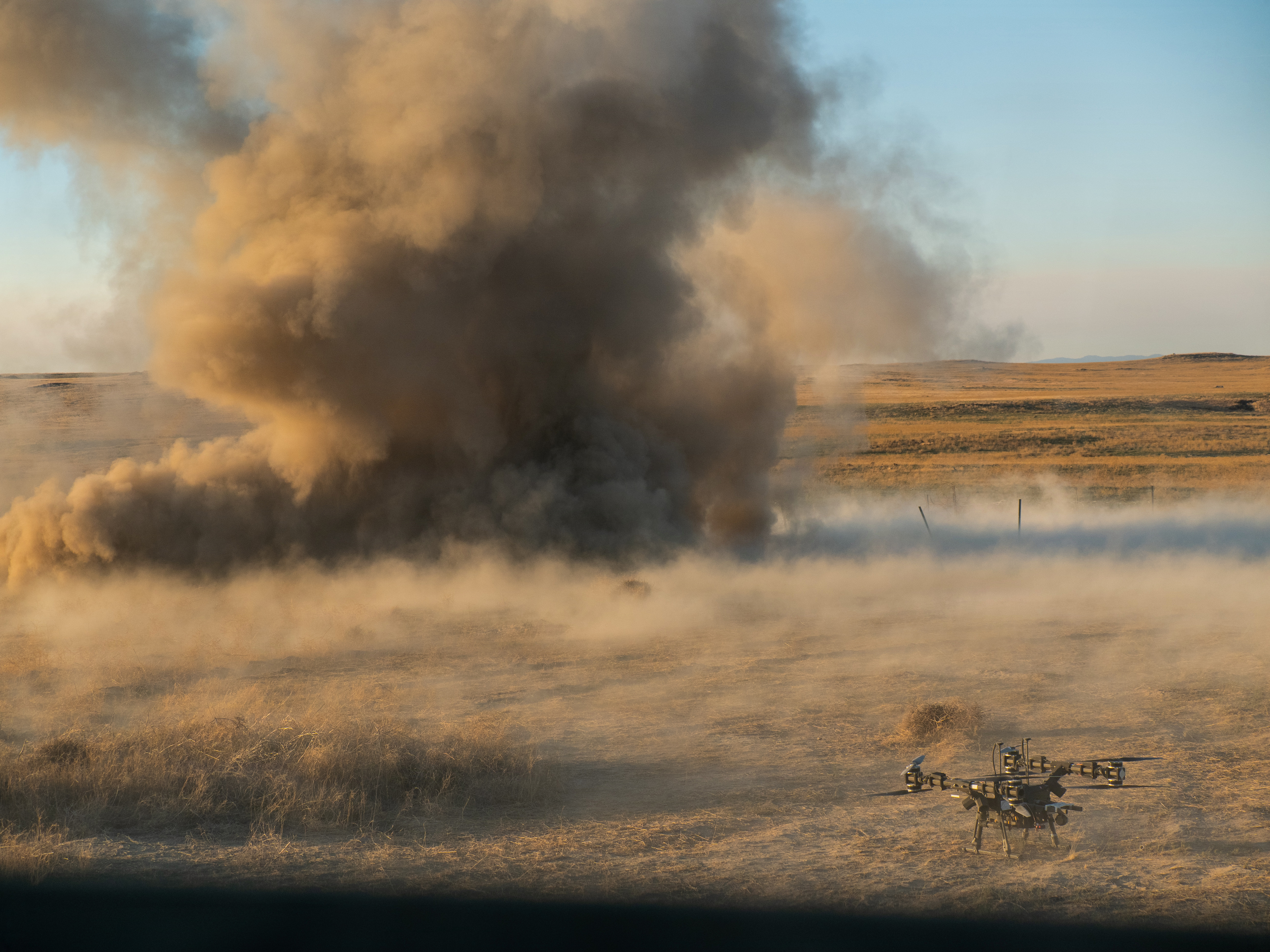 A Lorica Mule 28 unmanned aerial system rests on the ground as smoke rises in the background from the detonation of a live M1A3 Bangalore torpedo against a concertina wire obstacle. (Maj. W. Chris Clyne/Army National Guard)