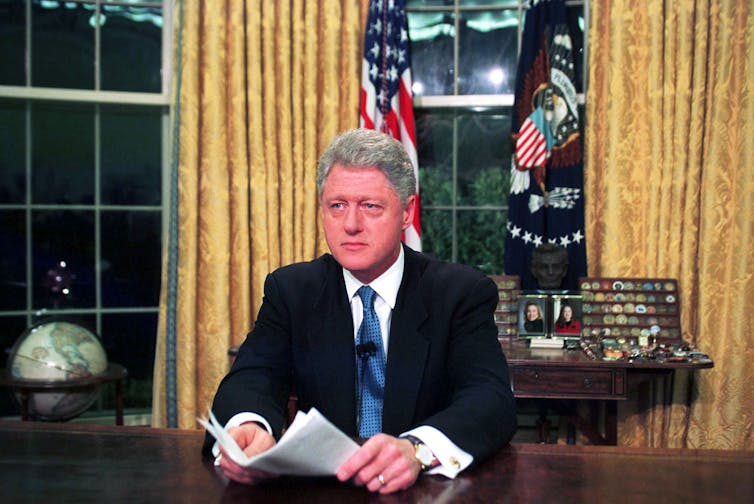 A gray-haired man in a dark jacket and blue tie, sitting at a desk in a very formal looking room.