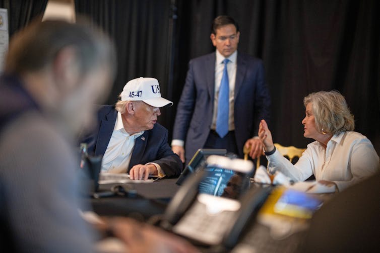 A man with a white hat and seated at a table listens to a woman speak to him.